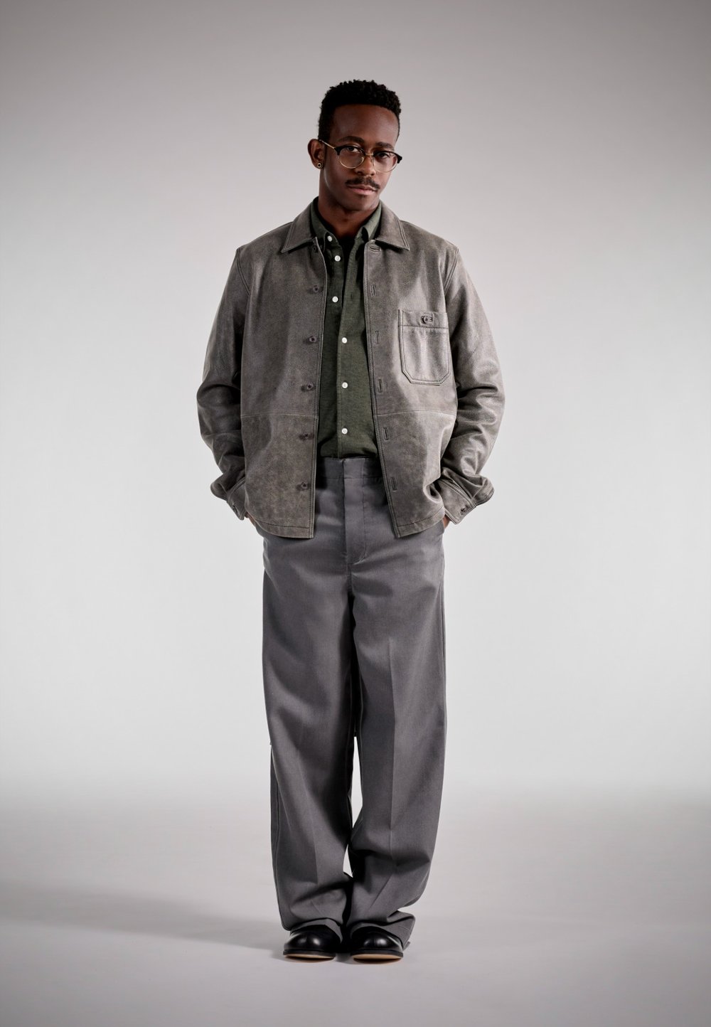 Giacca di pelle grigia sopra una camicia verde a bottoni, abbinata a pantaloni grigi larghi e scarpe nere; design semplice con tasca frontale.