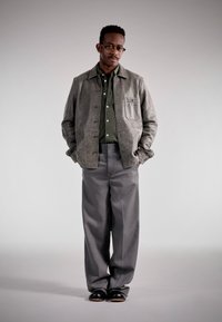 Giacca di pelle grigia sopra una camicia verde a bottoni, abbinata a pantaloni grigi larghi e scarpe nere; design semplice con tasca frontale.