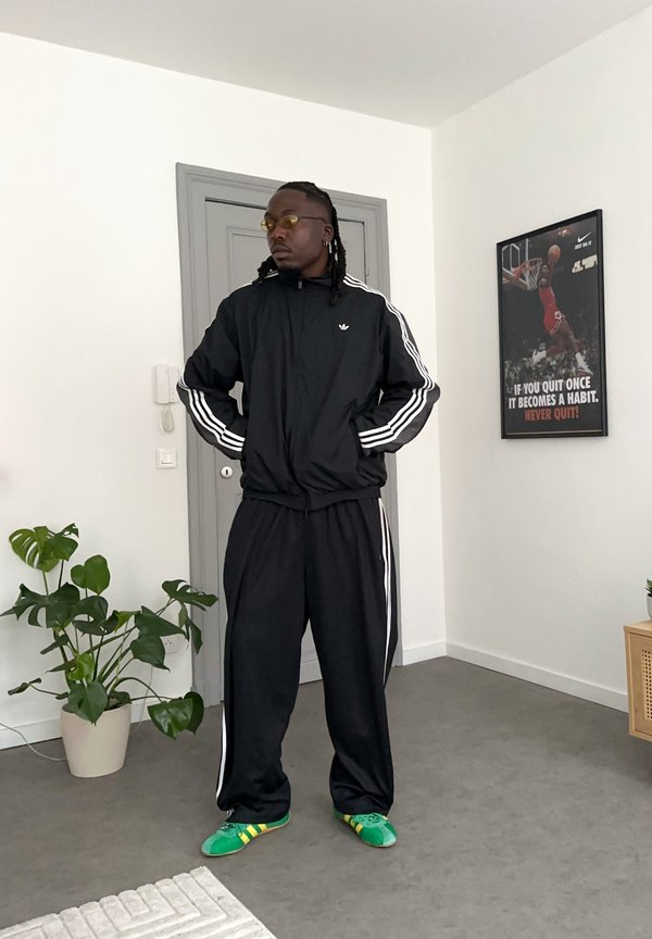 Juodas sportinis kostiumas su baltomis juostelėmis, žali sportbačiai su geltonais akcentais, vidaus aplinka su augalu ir sienų meno kūriniais fone.