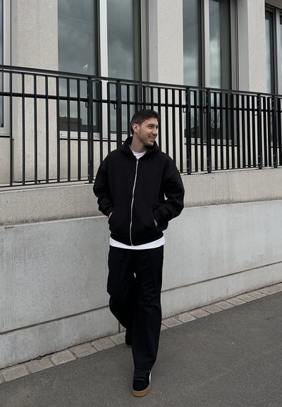 Joven caminando al aire libre junto a un edificio de concreto con grandes ventanales, vestido con sudadera negra con cremallera, pantalones negros y zapatillas deportivas.