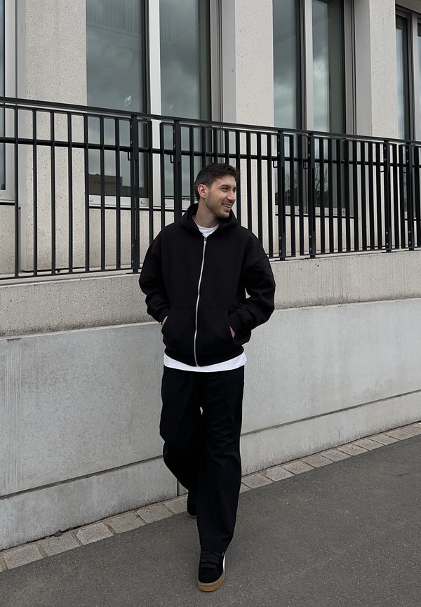 Młody mężczyzna idzie na zewnątrz obok betonowego budynku z dużymi oknami, ubrany w czarną bluzę z zamkiem, czarne spodnie i trampki.