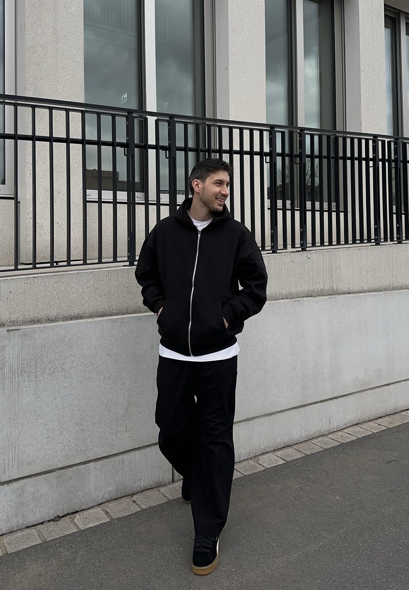 Noor mees kõndimas väljas betoonhoone kõrval, millel on suured aknad, kandes musta tõmblukuga kapuutsi, musti pükse ja tenniseid.