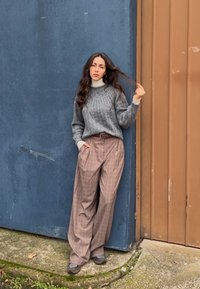 Femme debout contre un mur bleu et une porte marron, portant un pull gris, un pantalon large à motifs écossais, et des baskets grises, tenant ses cheveux d'une main.