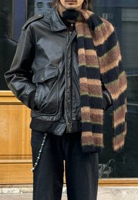 Veste bomber en cuir noir avec grandes poches, associée à une écharpe en laine rayée aux nuances de brun, vert et noir. Pantalon noir avec détail chaîne.