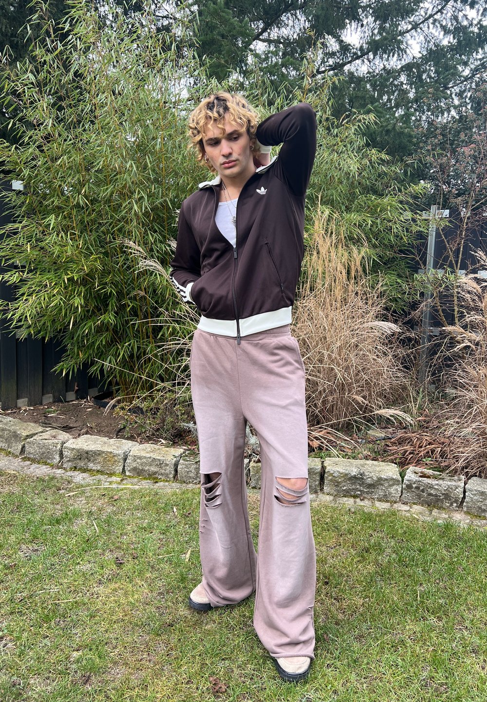 Veste de survêtement marron à fermeture éclair avec accents blancs ; t-shirt gris clair en dessous. Pantalon large rose pâle avec de grandes déchirures aux genoux.