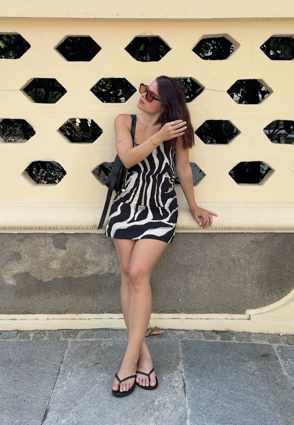 Femme portant des lunettes de soleil et une robe à imprimé zèbre, appuyée contre un mur crème avec des ouvertures géométriques, ajustant ses cheveux.