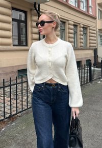 Femme portant un cardigan blanc boutonné, un jean foncé, des lunettes de soleil noires et tenant un sac à main noir, marchant sur un trottoir en ville.