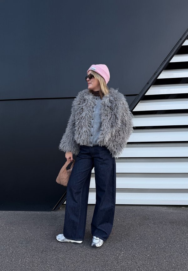 Femme portant une veste grise duveteuse, un bonnet rose, des lunettes de soleil, un jean large et des baskets, tenant un sac tissé à côté d'un escalier moderne noir et blanc.