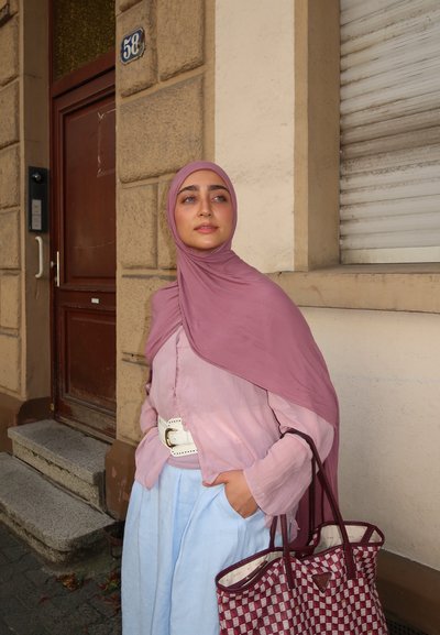 Camisa de manga larga rosa claro con un cinturón blanco, combinada con pantalones anchos azul claro. Un pañuelo de cabeza rosa intenso y un bolso a cuadros burdeos.