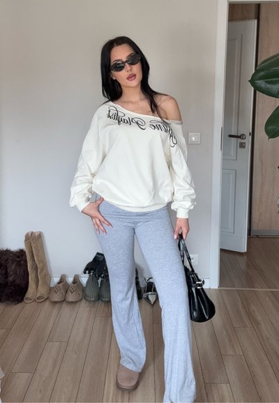Mujer con gafas de sol, sudadera blanca con los hombros descubiertos, pantalones grises, sosteniendo un bolso negro, de pie en una habitación con zapatos en el suelo.