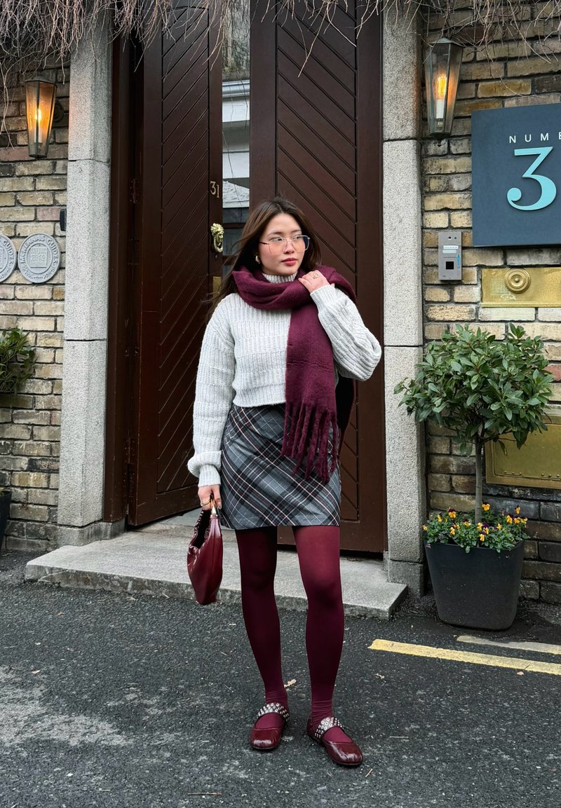 Frau mit Brille, die einen grauen Pullover, einen karierten Rock, einen bordeauxroten Schal, Strumpfhosen und Schuhe trägt, hält eine bordeauxrote Tasche und steht vor einer Tür eines Backsteingebäudes.