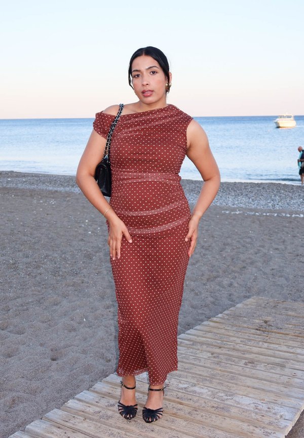 Una mujer con un vestido burdeos de lunares y zapatos negros de tacón posa en una pasarela junto a la playa, con el mar y un barco detrás.