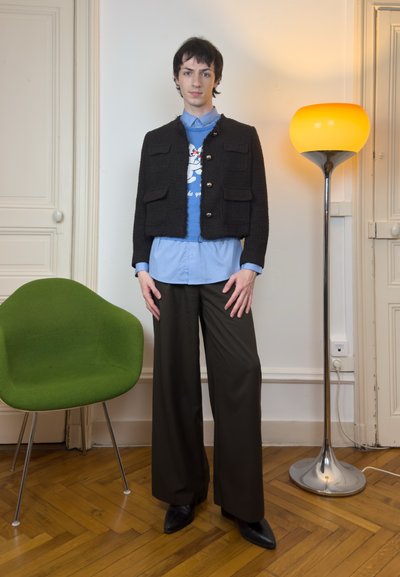 Persona joven de pie en interiores con chaqueta negra, camisa azul con gráfico de dibujos animados, pantalones anchos negros, cerca de una silla verde y una lámpara de pie amarilla.