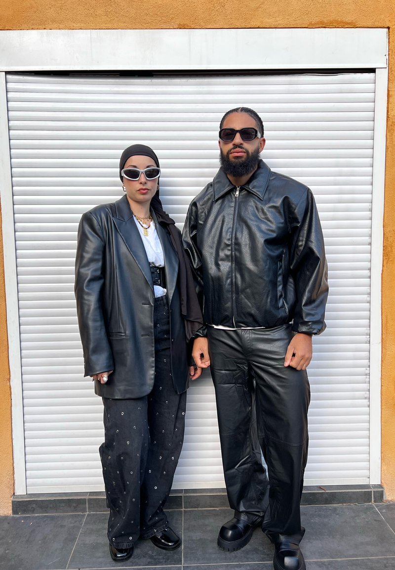 Deux personnes posent devant une porte de garage blanche à enroulement. La femme porte un blazer en cuir noir et un pantalon, avec un haut blanc et des lunettes de soleil. L'homme arbore une veste zippée en cuir noir et un pantalon, avec des chaussures noires épaisses.