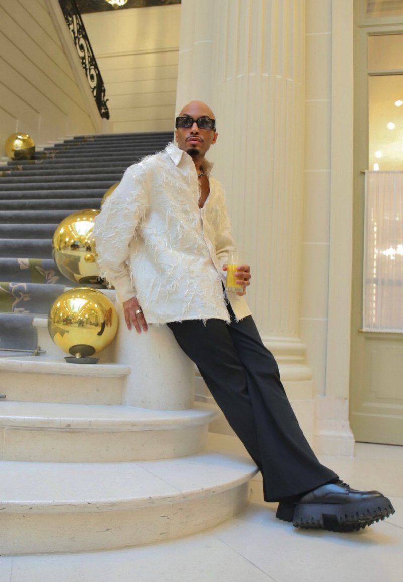 Homme portant une chemise blanche texturée, un pantalon noir et des chaussures épaisses, appuyé contre une colonne près d'un escalier décoré de grandes sphères dorées réfléchissantes.