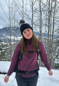Femme portant un bonnet à pompons noir et blanc et une veste polaire violette, avec un sac à dos noir, entourée d'arbres enneigés et de montagnes.