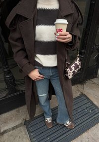 Personne portant un manteau marron, un pull rayé et un jean déchiré, tenant une tasse de café et un sac à main à imprimé vache devant un bâtiment.
