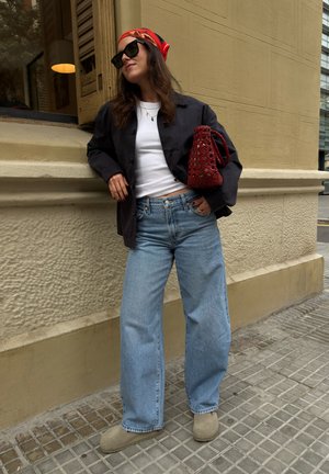 Mujer con gafas de sol, pañuelo rojo en la cabeza, chaqueta negra, camisa blanca, vaqueros azules, zapatos marrones claros, sosteniendo un bolso rojo tejido, apoyada en una pared beige al aire libre.
