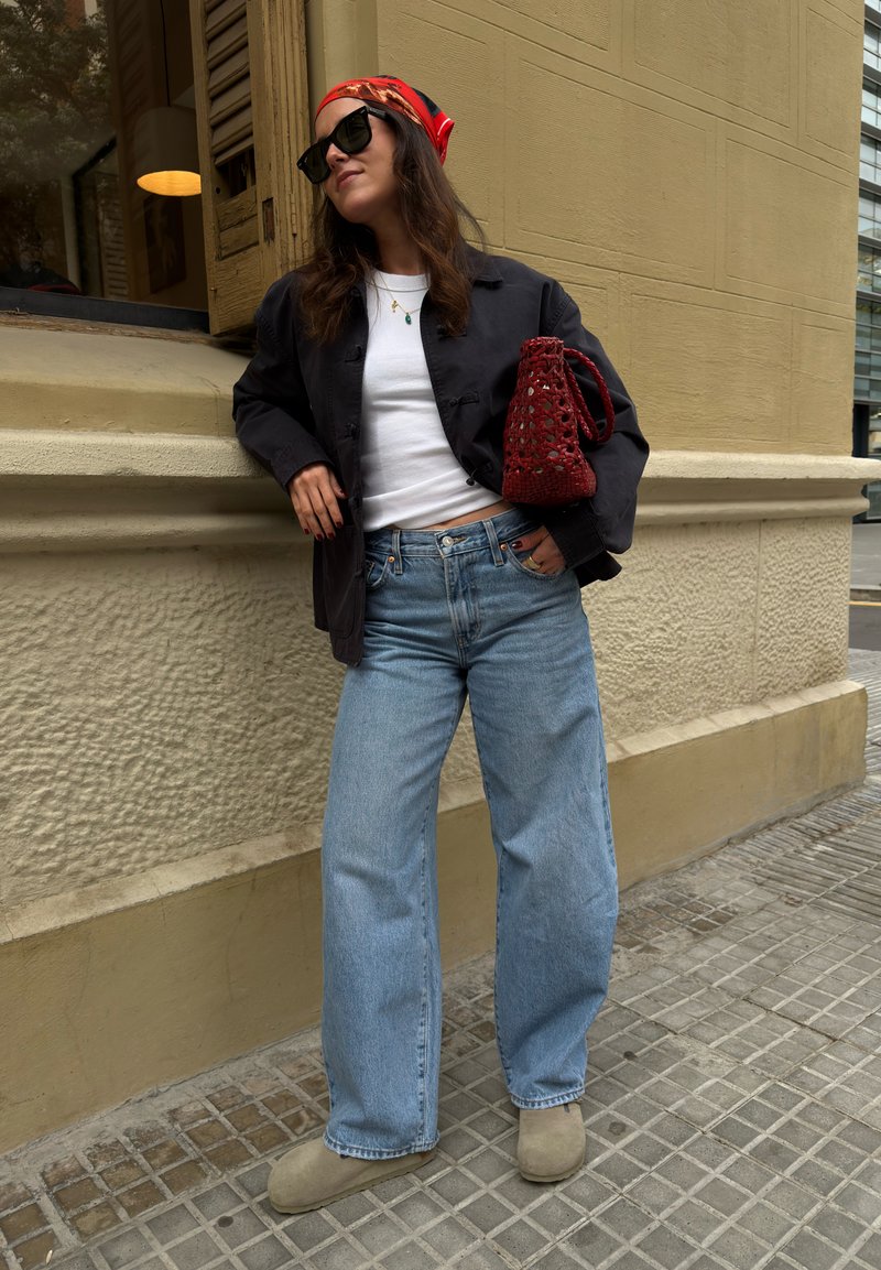 Femme portant des lunettes de soleil, foulard rouge, veste noire, chemise blanche, jean bleu, chaussures couleur sable, tenant un sac tissé rouge, appuyée contre un mur beige à l'extérieur.