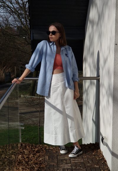 Camisa de botón azul claro con logo, top corto coral, falda maxi blanca y zapatillas negras. Entorno al aire libre con vegetación y hojas.