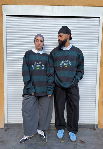 Dos personas con camisetas a rayas a juego que dicen "Originals Dept 1972" están paradas frente a una persiana blanca; una lleva pantalones anchos grises y la otra pantalones a rayas finas.