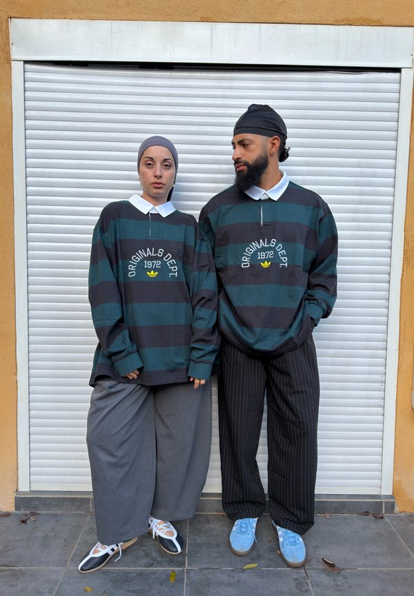 Zwei Personen tragen passende gestreifte "Originals Dept 1972"-Shirts und stehen vor einem weißen Rollladen, eine in weiten grauen Hosen, die andere in Nadelstreifenhosen.