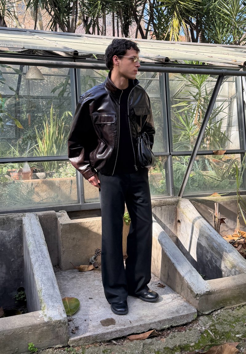 Homme portant une veste en cuir foncé, un pantalon noir et des bottes se tient à l'intérieur d'une serre avec des plantes et des plates-bandes en béton.