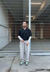 Schwarzes kurzärmliges Hemd mit Knöpfen über einem grauen Langarmshirt, kombiniert mit hellgrauen Hosen und schwarzen Sneakers mit grünen Akzenten.