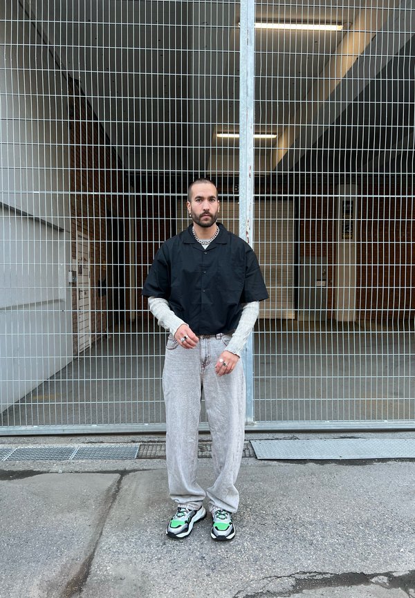 Schwarzes kurzärmliges Hemd mit Knöpfen über einem grauen Langarmshirt, kombiniert mit hellgrauen Hosen und schwarzen Sneakers mit grünen Akzenten.