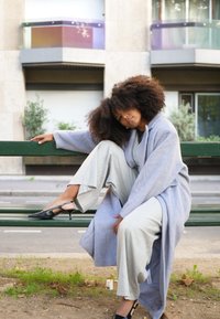 Ανοιχτό γκρι υπερμεγέθη ρόμπα, γκρι παντελόνια φαρδιάς γραμμής και μαύρες τακούνια. Σγουρά μαλλιά, καθισμένη σε ένα πράσινο παγκάκι σε αστικό περιβάλλον.