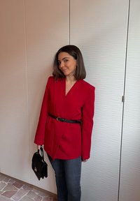 Donna con capelli castani fino alle spalle, che indossa un blazer rosso con cintura, jeans scuri e tiene una borsa nera, in piedi davanti a pannelli murali testurizzati.