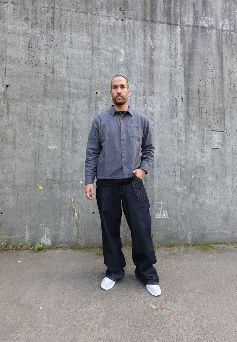 Chemise grise à boutons avec une poche à l'avant, portée sur un top noir. Pantalons en denim foncé ample et baskets blanches sur un fond en béton.