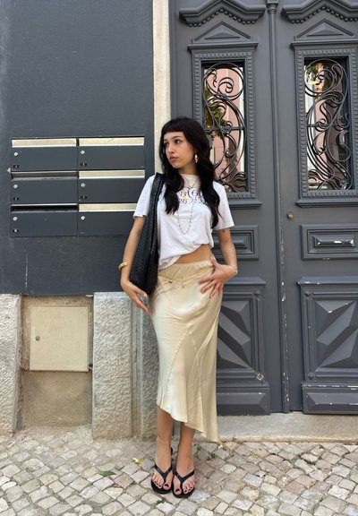Mujer con el pelo largo y oscuro vestida con camiseta blanca, falda de satén color crema, sandalias negras y joyas de oro, de pie junto a una puerta decorativa oscura y buzones.