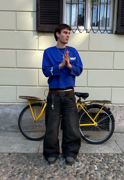 Top azul recortado de manga larga, vaqueros negros de gran tamaño y zapatos oscuros. Bicicleta amarilla en el fondo, con un asiento de madera y acentos negros.