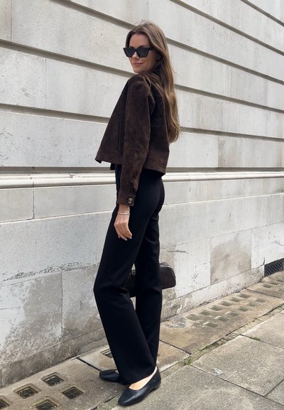 Chaqueta corta de ante marrón, pantalones de pierna ancha negros, bailarinas negras y un pequeño bolso, con el pelo liso y gafas de sol oscuras, frente a una pared de piedra.