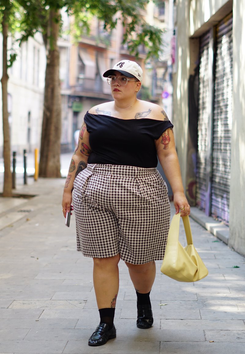 Top noir à épaules dénudées, short à carreaux taille haute et sac jaune. Lunettes rondes, casquette de baseball blanche et mocassins noirs. Cadre urbain.