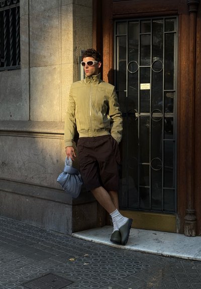 Chaqueta bomber verde claro, pantalones cortos marrones, calcetines blancos, zapatos verde oliva y una bolsa azul. El modelo se apoya contra una pared de piedra.