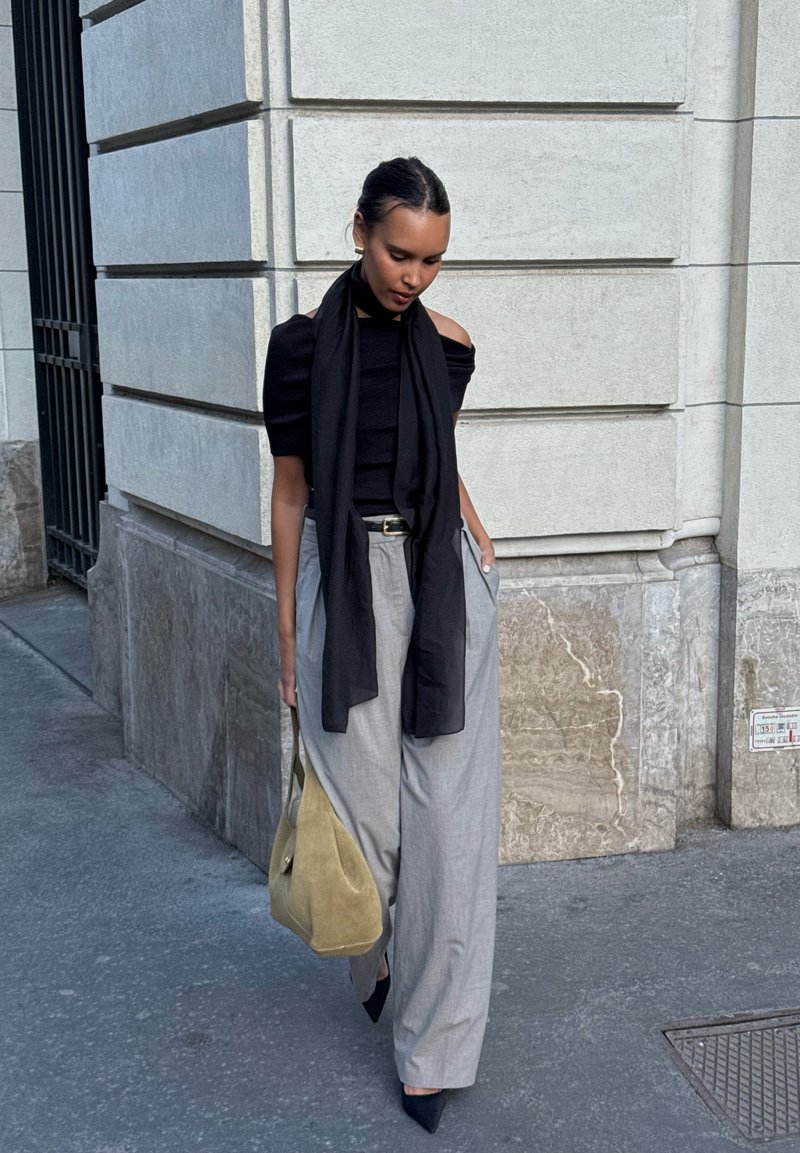 Femme marchant sur le trottoir de la ville portant un haut noir avec une écharpe, un pantalon large gris, et portant un grand sac à main beige.