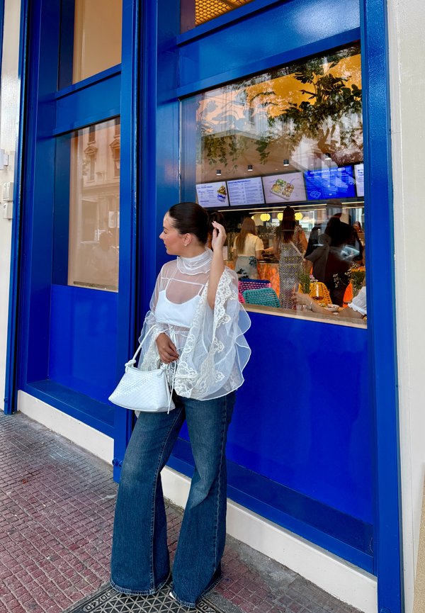 Femme en blouse blanche en dentelle et jean bleu se tient devant une fenêtre encadrée de bleu d'un restaurant animé avec des personnes à l'intérieur et des écrans de menu visibles.