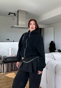 Μαύρος κοντός ζακέτας με φερμουάρ, χαλαρή εφαρμογή, σε συνδυασμό με μαύρη φούστα skater και αλυσίδα στη μέση, συμπληρωμένο με φαρδιά μαύρα τζιν.
