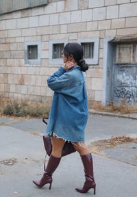 Džinsa oversized krekls ar saplēstu apakšu, kombinēts ar spīdīgām burgundas augstiem zābakiem virs ceļgala un saskaņotu somiņu. Fona pelēks akmens siena.