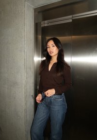 Rjava dolga športna srajca z gumbi, svetlo modri hlače z visokim pasom. Stoječa spredaj pred kovinskimi dvigali s betonskimi stenami, drži očala.