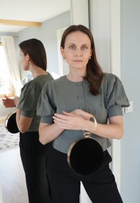 Apvalus juodas delninukas su auksinėmis detalėmis. Trumpomis rankovėmis pilka palaidinė su faktūruota medžiaga ir pūstomis rankovėmis. Atspindintis veidrodinis fonas.