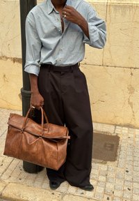 Hnedá kožená taška typu duffle s hornými rúčkami a zipsom, v kombinácii so sivou košeľou na gombíky a čiernymi širokými nohavicami.