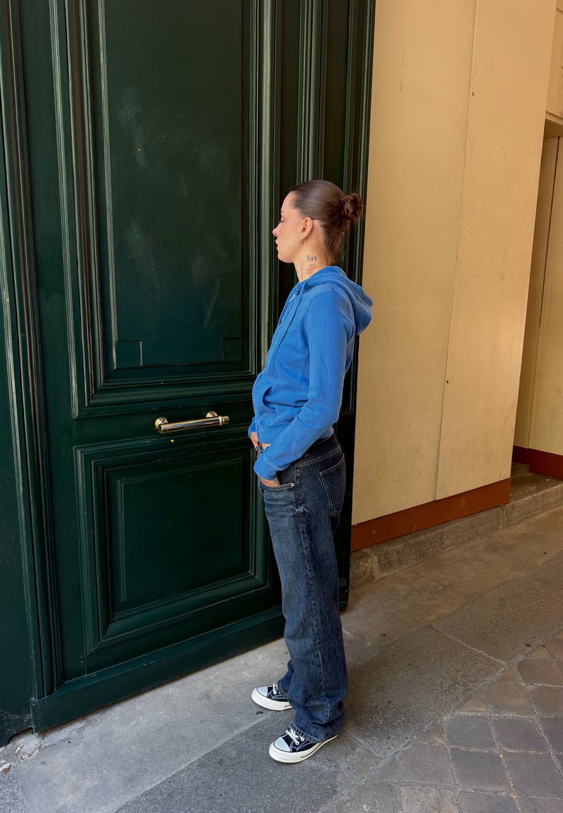 Jeune femme avec un chignon, portant un sweat à capuche bleu et un jean, debout les mains dans les poches, face à une grande porte vert foncé dans un couloir extérieur.