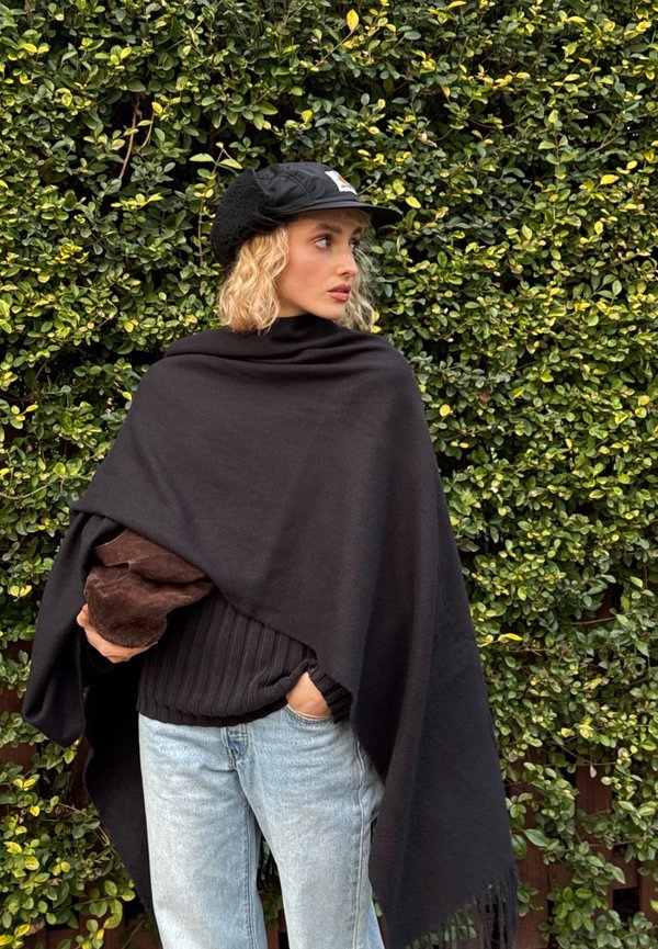 Zwarte poncho met een gladde textuur en franjes aan de randen, gedragen over een geribbelde zwarte trui. Model houdt een bruin pluchen voorwerp vast tegen een onscherpe groene achtergrond.