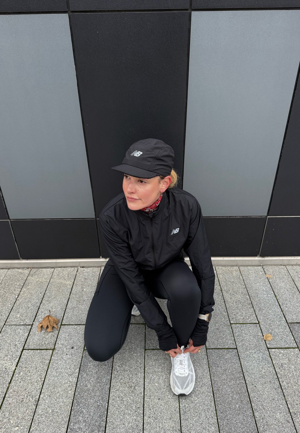 Veste et pantalon de sport noirs, baskets blanches, casquette noire, en position accroupie, surface en béton, fond géométrique gris.
