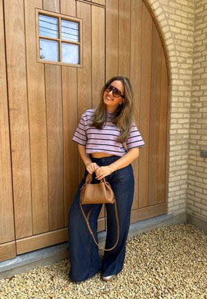 Top corto morado a rayas, pantalones de pierna ancha oscuros y un bolso marrón. De pie contra una pared de madera con una pequeña ventana. Suelo de piedras texturizadas.