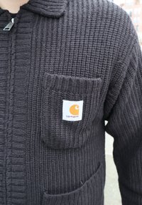 Κοντινό πλάνο ενός ατόμου που φοράει σκουρό καφέ πλεκτή ζακέτα με φερμουάρ και τσέπη στο στήθος με λευκή ετικέτα λογότυπου Carhartt.