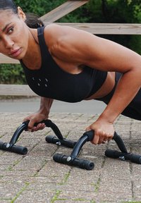 Černé opěrky na kliky s texturovanými úchopy a gumovými konci, umístěné na kamenném povrchu. Atlet ve černém sportovním oblečení předvádí jejich použití.